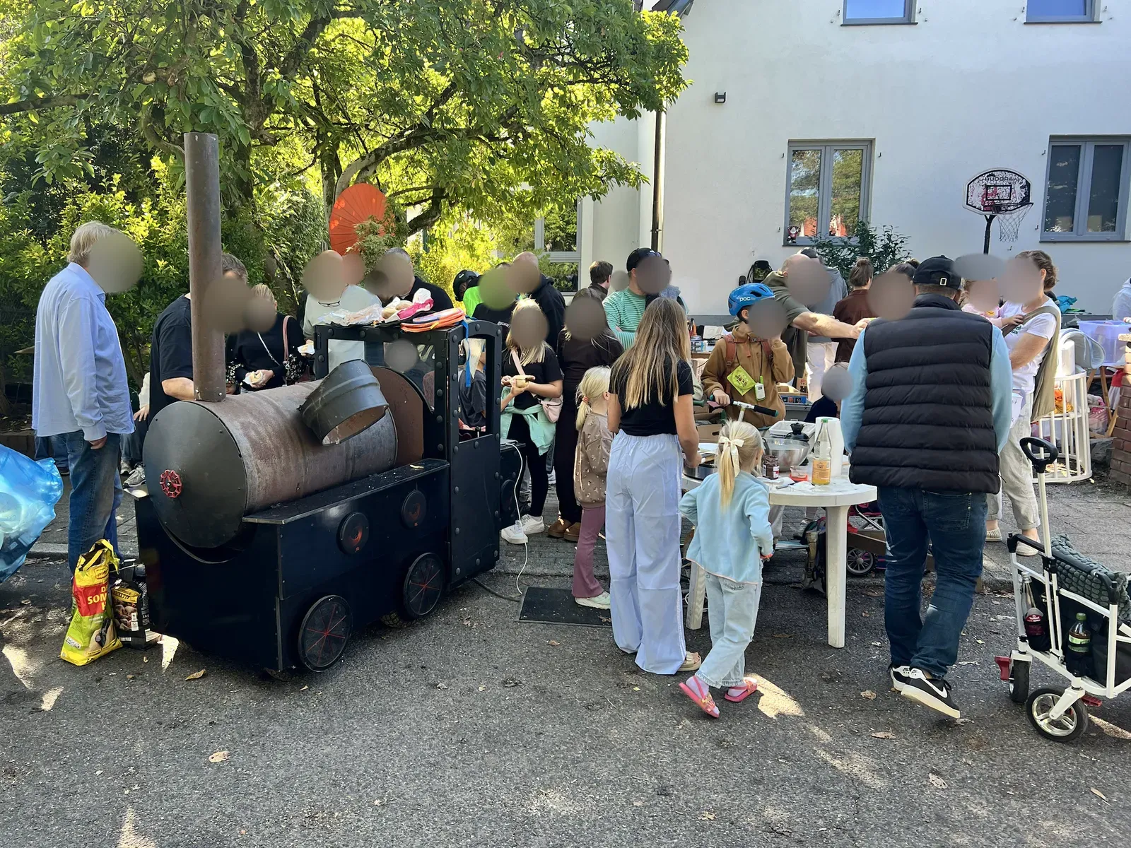 Holzkohlegrill in Form einer schwarzen Dampflok am Straßenrand, drumherum viele Erwachsene und Kinder