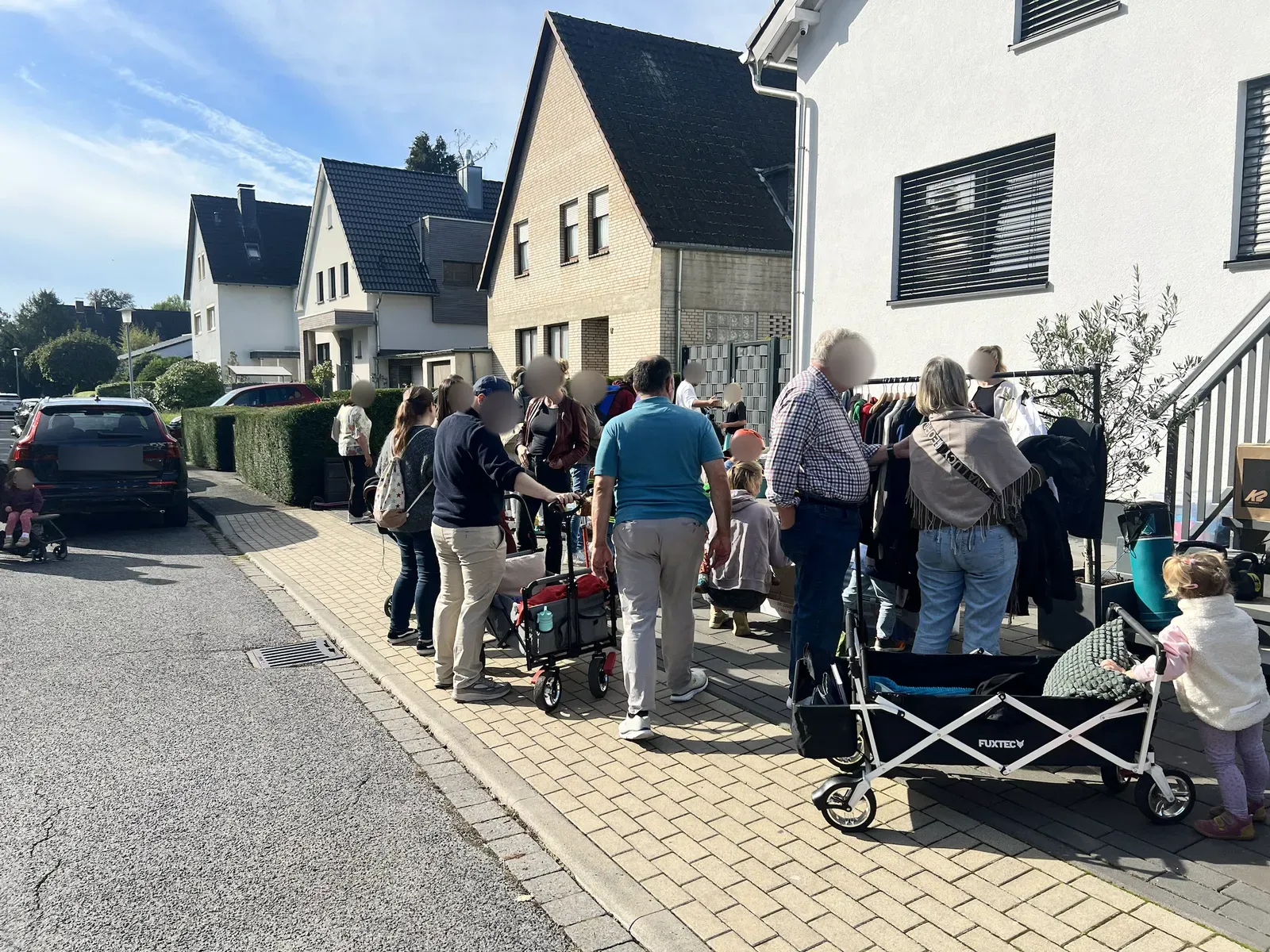 Viele Leute an einer Kleiderstange auf einem Gehweg vor Wohnhäusern, davor zwei Bollerwagen