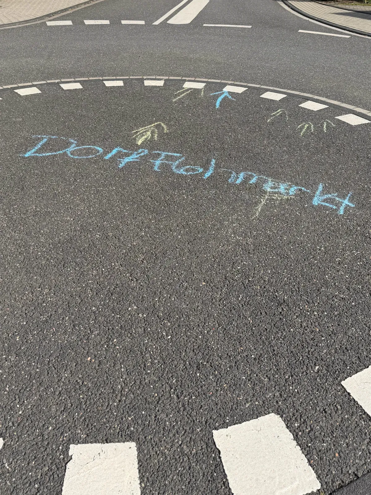 Mit blauer Kreide auf die Straße geschrieben: „Dorfflohmarkt“, dazu gelbe und blaue Kreidepfeile auf einer Verkehrsinsel