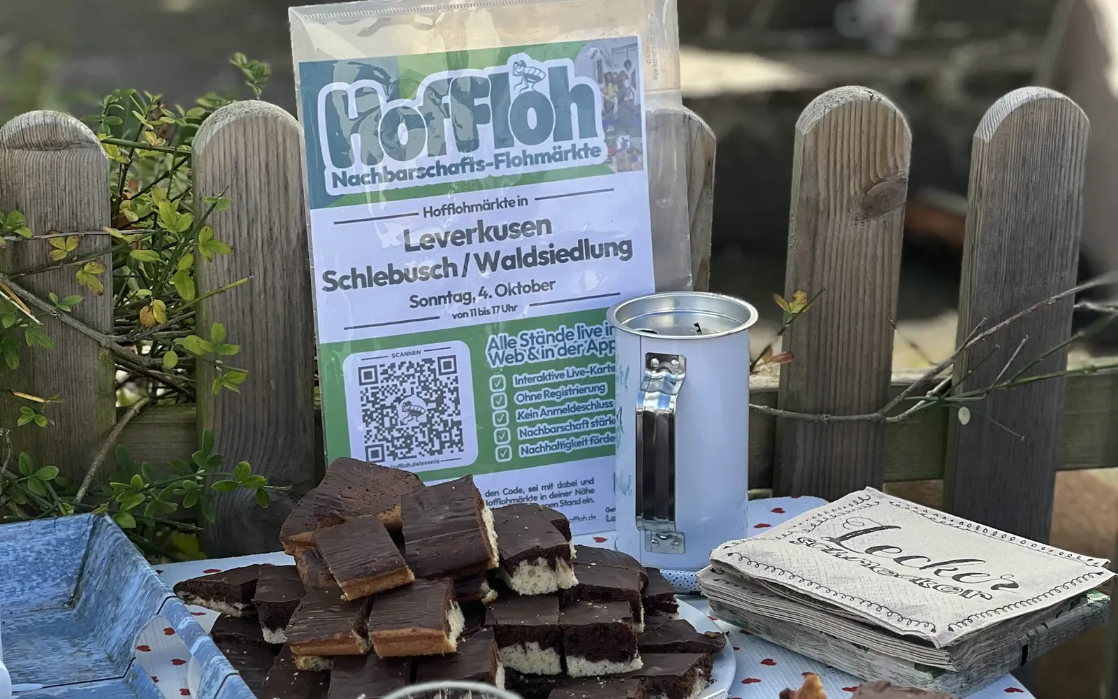 HofFloh-Plakat „Leverkusen Schlebusch/Waldsiedlung, Sonntag, 4. Oktober“ an einem Holzzaun, davor ein Teller mit Schokokuchen und ein Stapel Servietten