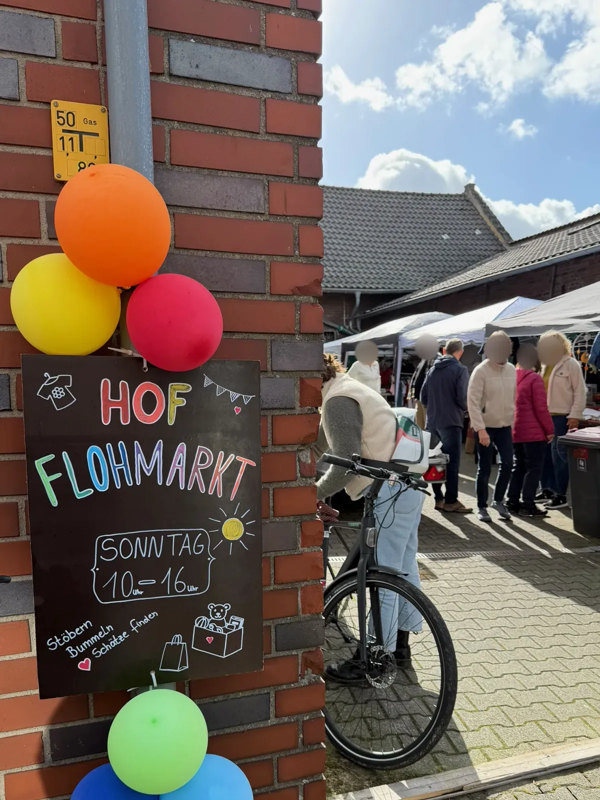 Kreidetafel „Hof Flohmarkt, Sonntag 10 bis 16 Uhr, Stöbern, Bummeln, Schätze finden“ mit bunten Luftballons an einer Backsteinwand, dahinter Besucher an Pavillons in einem Innenhof