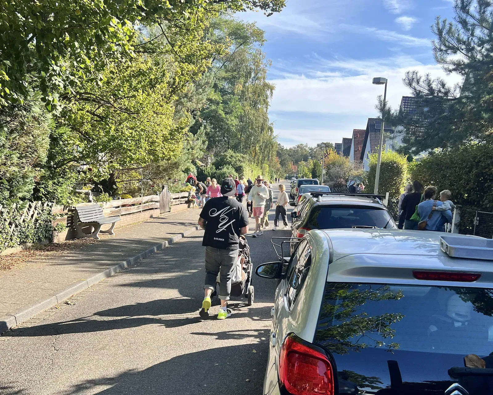 Sonnige Wohnstraße mit parkenden Autos, ein Mann schiebt einen Kinderwagen, weiter hinten viele Leute zu Fuß